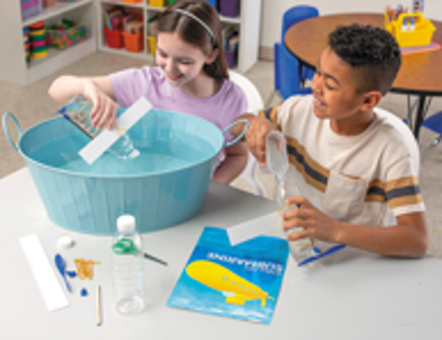 Two children playing with a science experiment kit floating submarine on a table in a classroom setting.