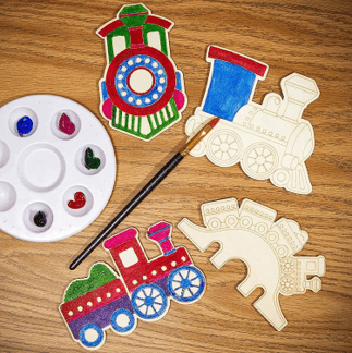Children's train-themed craft set with paint, brushes, and unpainted train pieces on a wooden surface.